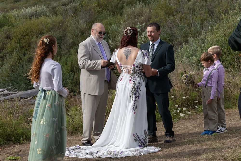 Outdoor wedding ceremony with bride in floral white gown, groom, officiant, and guests gathered in a natural hillside setting.