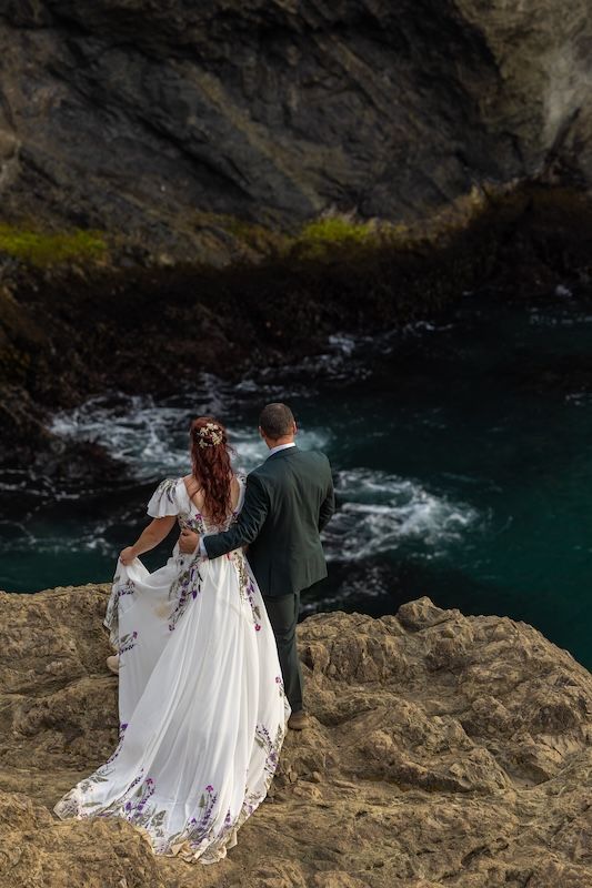 Bride and groom stand on rocky coastal cliffs overlooking turquoise ocean waters during their outdoor wedding ceremony.