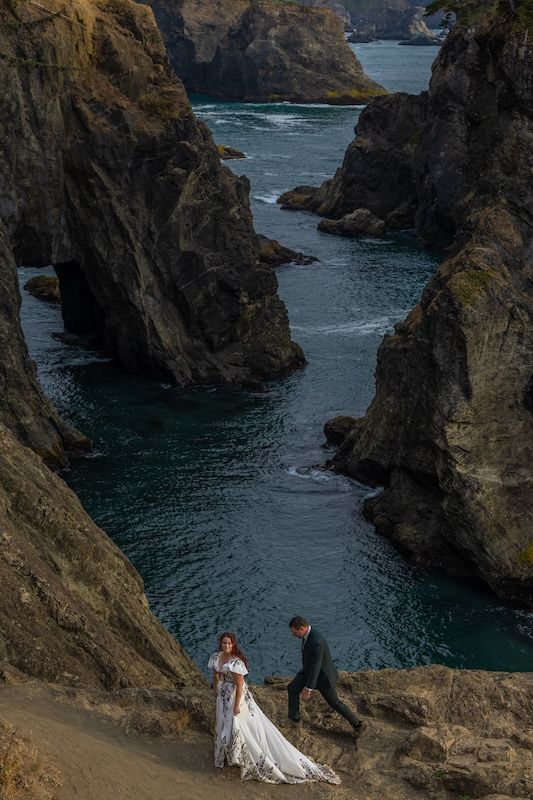 Bride and groom stand on rocky coastal cliffs with dramatic sea stacks and turquoise water below.