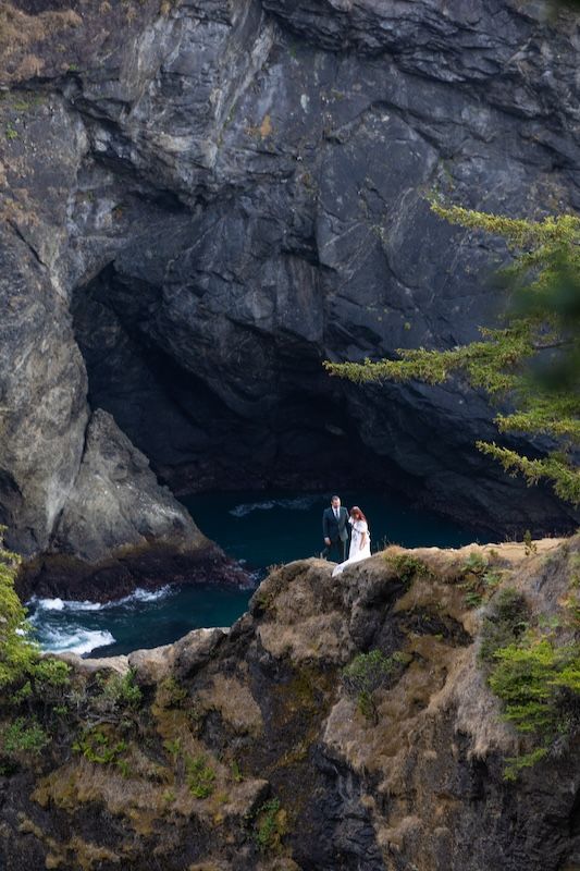 Bride and groom stand on rocky coastal cliff with sea cave and turquoise water backdrop during outdoor wedding ceremony.