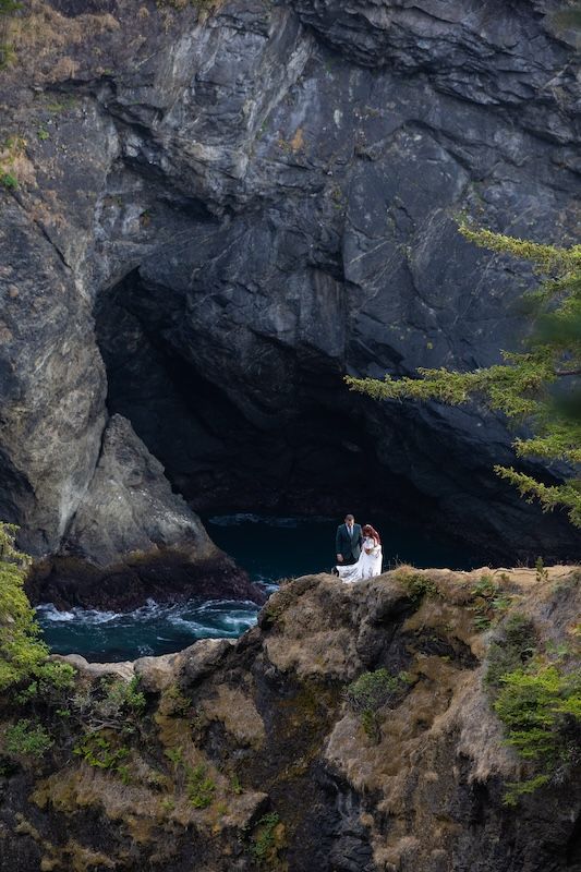 Bride and groom stand on rocky coastal cliffs beside a dramatic sea cave with ocean waves below.