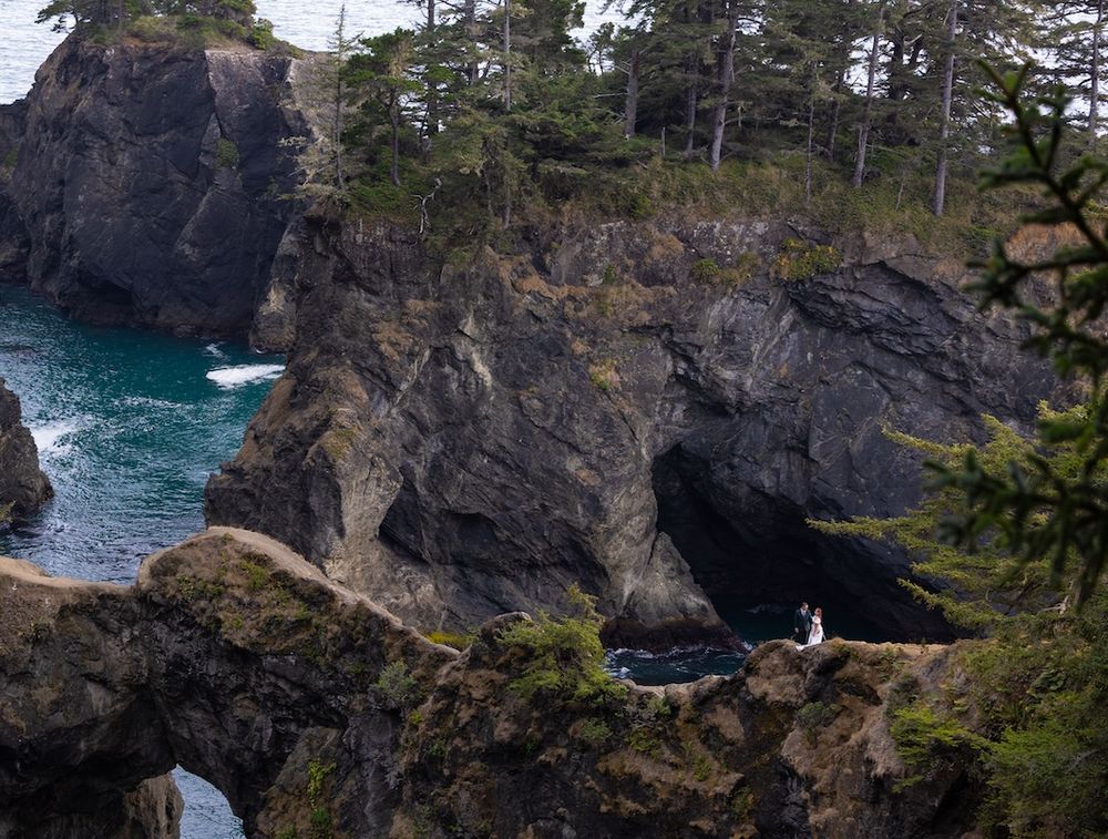 Couple in wedding attire standing on rocky coastal cliffs with sea caves and turquoise water below.