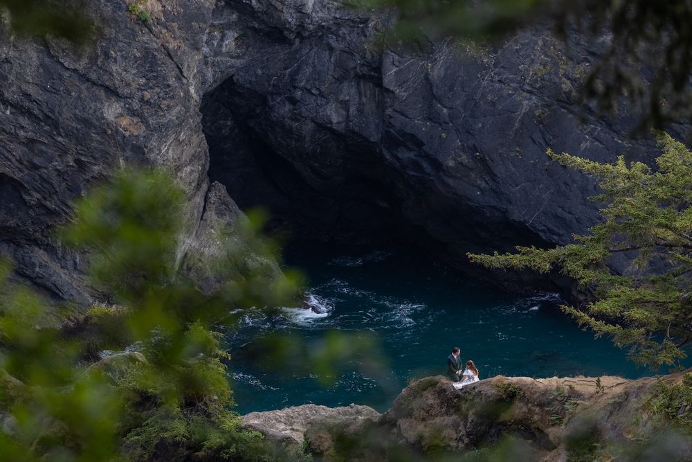Person sitting on rocks beside a turquoise sea cave with dark cliffs and lush green trees framing the scene.