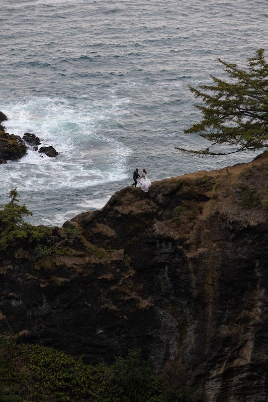 Couple standing on a rocky coastal cliff edge overlooking crashing ocean waves with a evergreen tree nearby.