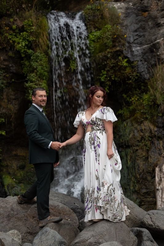 Couple in wedding attire holding hands before a waterfall, bride in floral gown, groom in dark suit, standing on rocks.