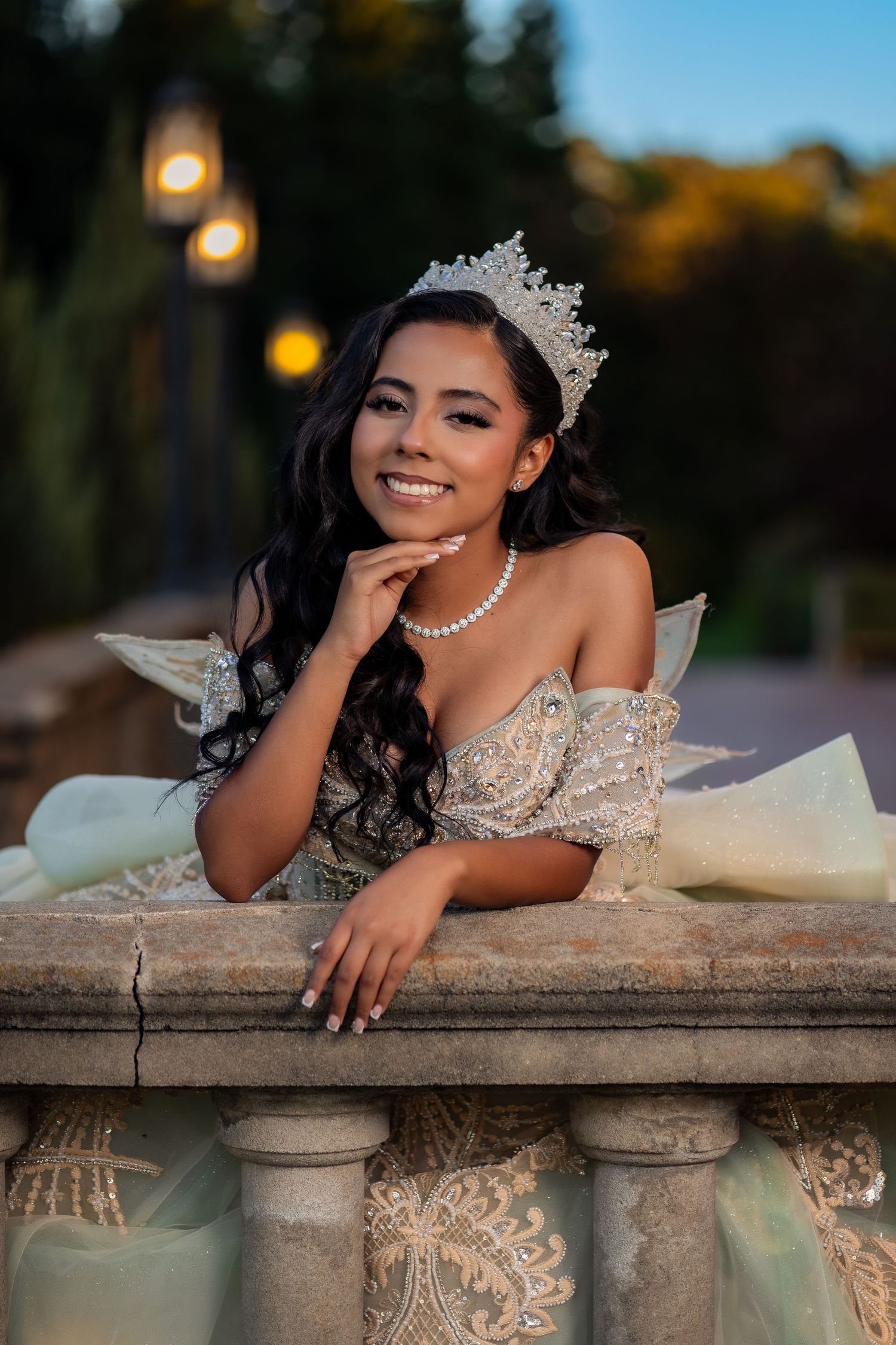 Young Latina woman in elegant lace gown and crystal tiara smiling at outdoor quinceañera celebration.