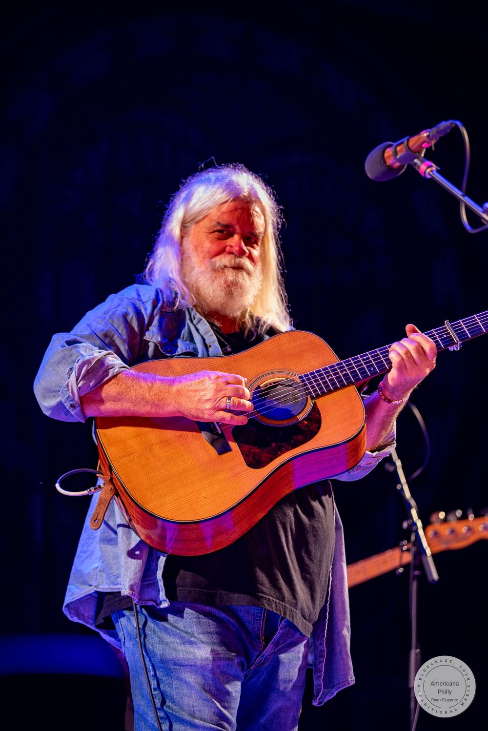 Elderly musician with long white hair and beard playing acoustic guitar on stage under colorful lights.