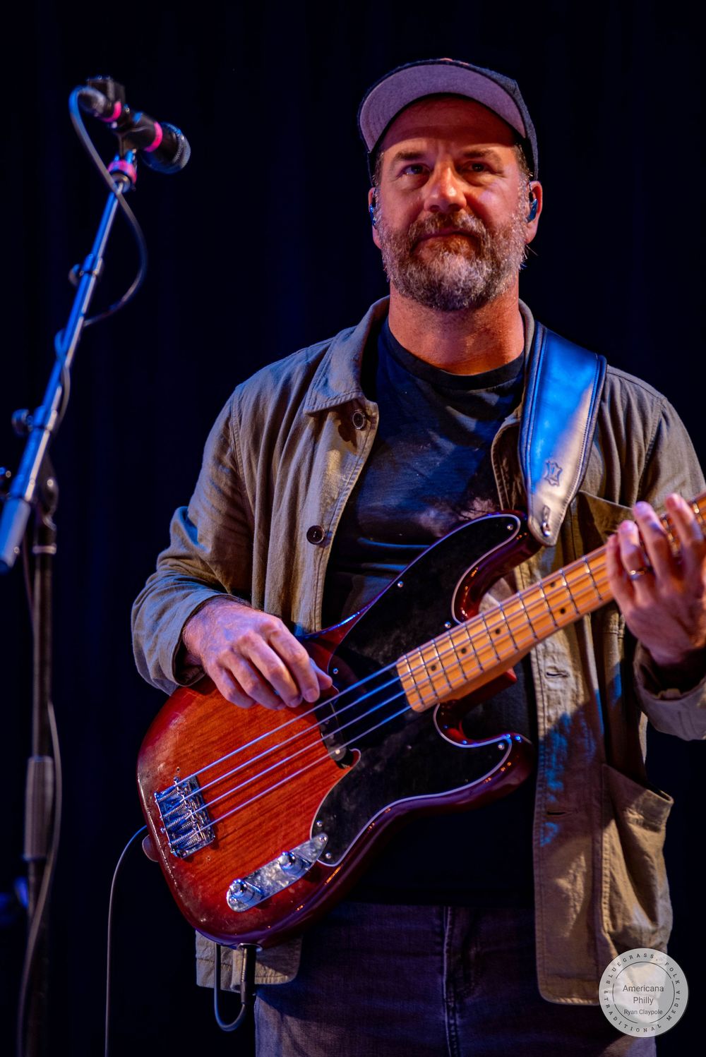 Bearded male bassist performing on stage with a red electric bass guitar and cap under blue stage lighting.