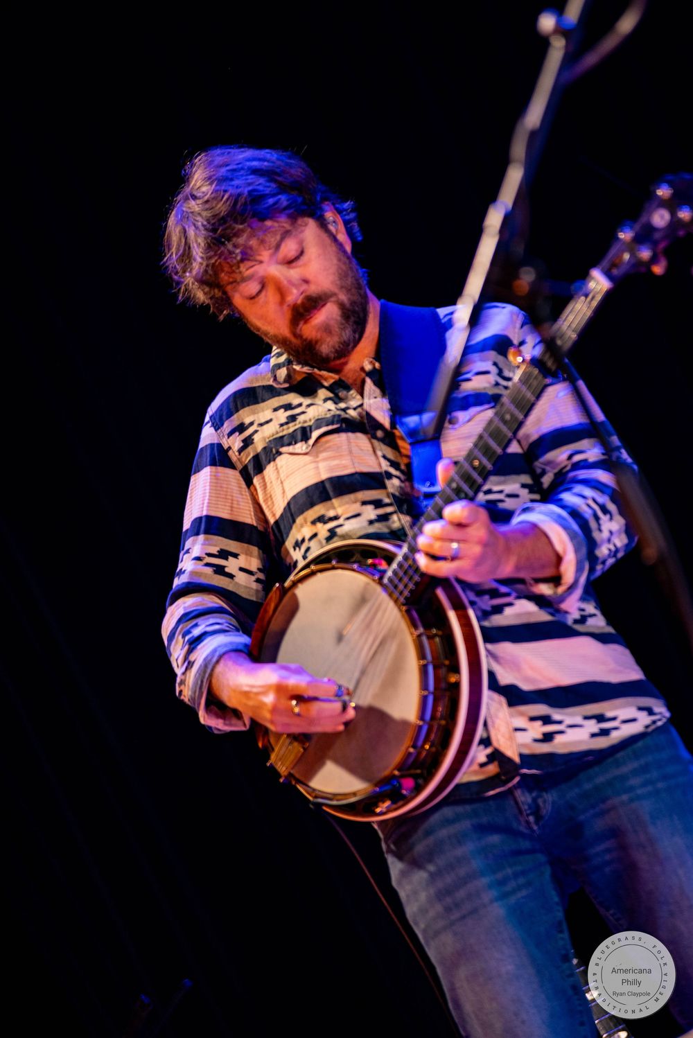 Bearded musician playing a banjo on stage, wearing a striped shirt under blue stage lighting.