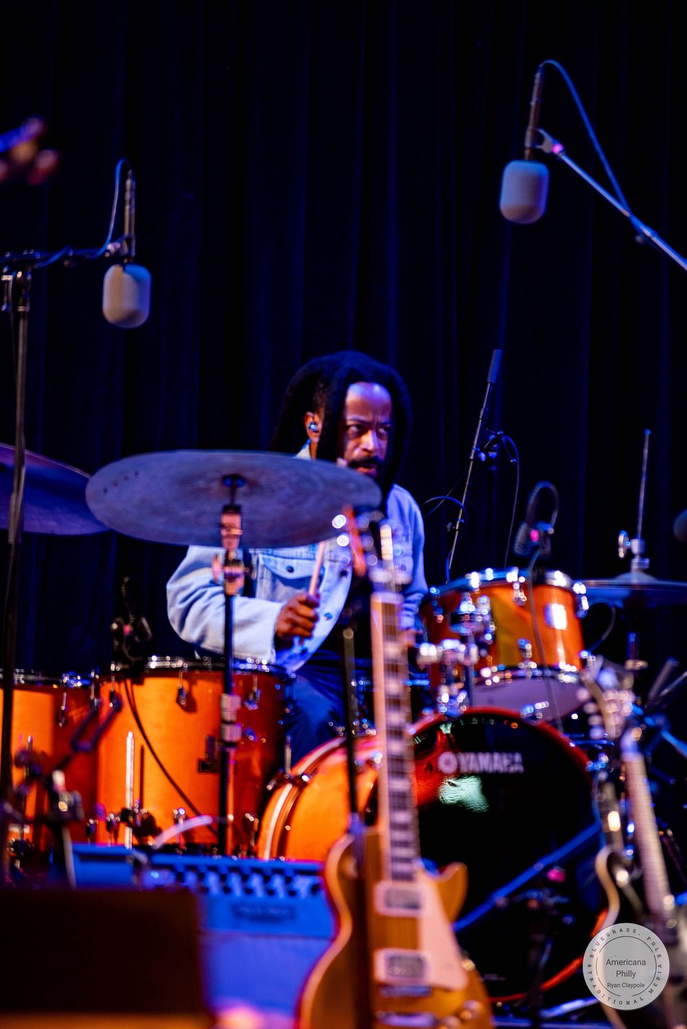 A Black musician plays drums on a Yamaha kit with cymbals on a dimly lit stage, with a guitar visible in the foreground.
