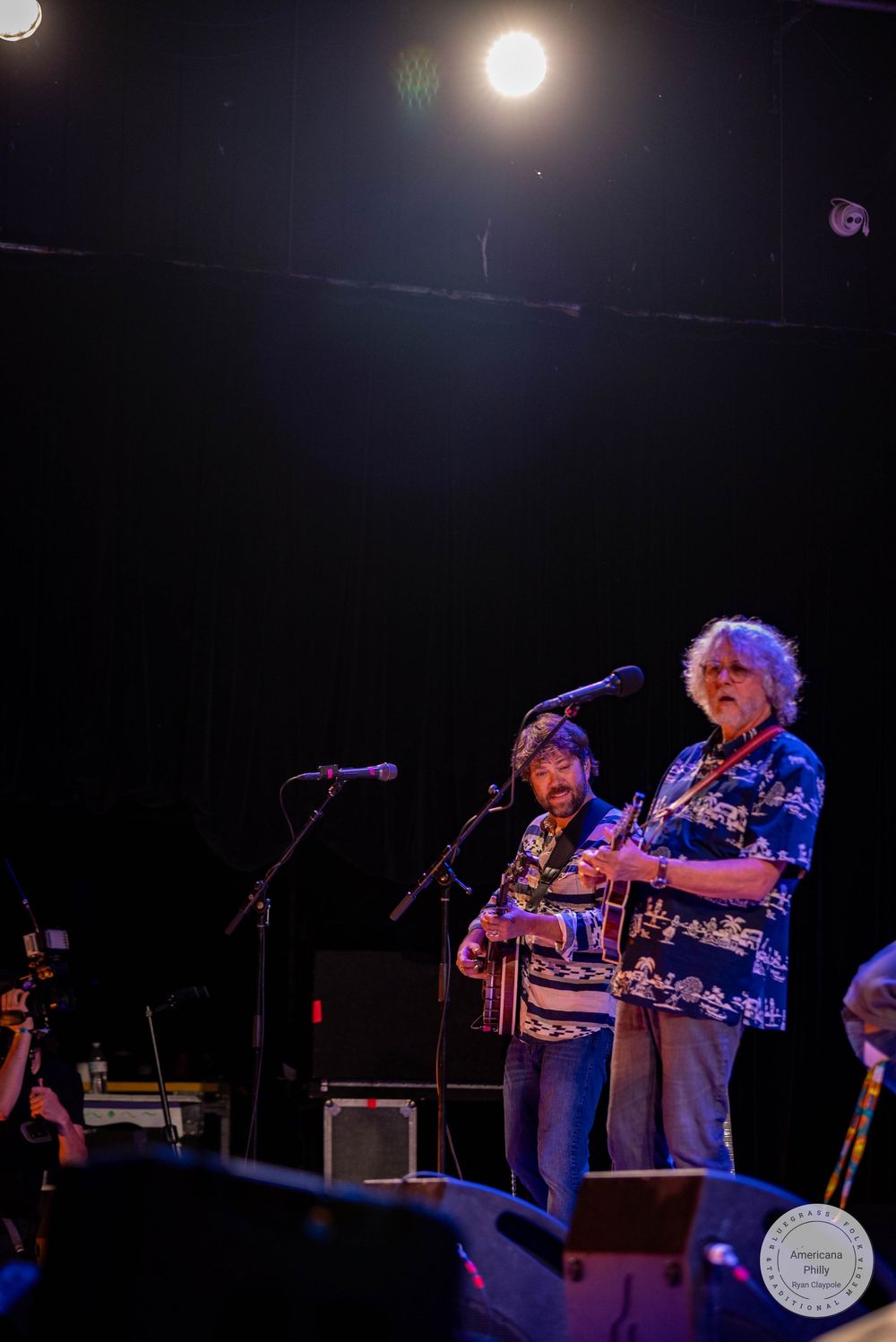 Two musicians perform on a dimly lit stage with microphones and instruments at a live concert.