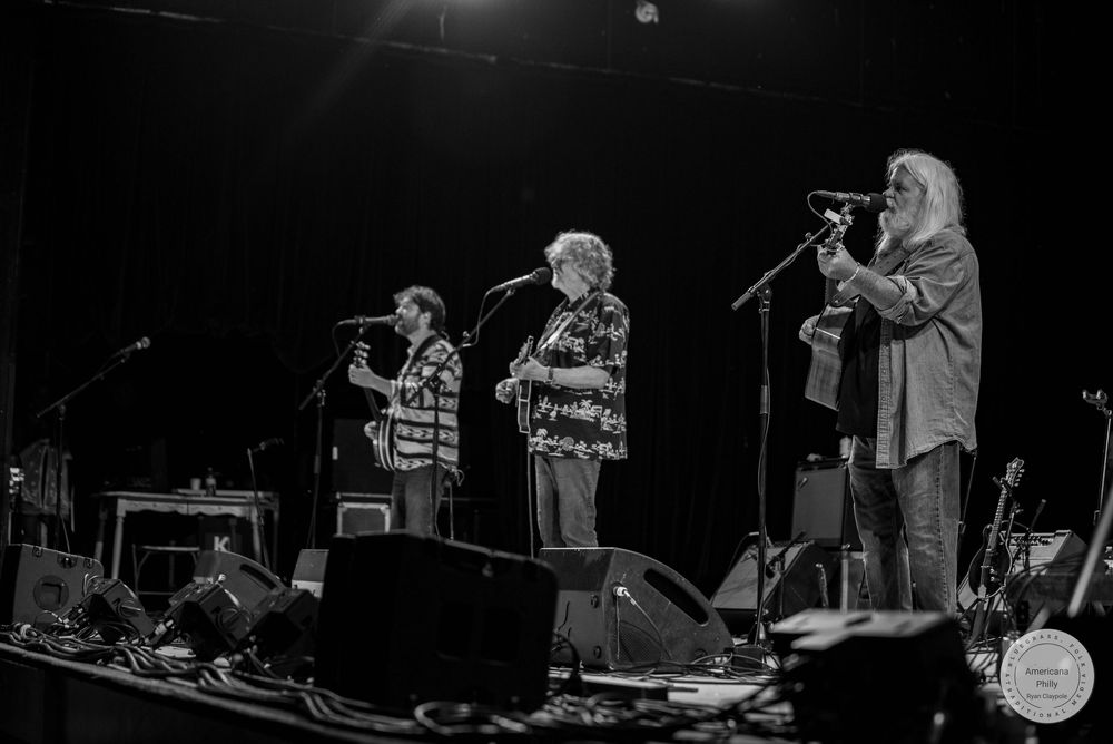 Three musicians performing on stage with microphones and guitars in a black and white concert photo.
