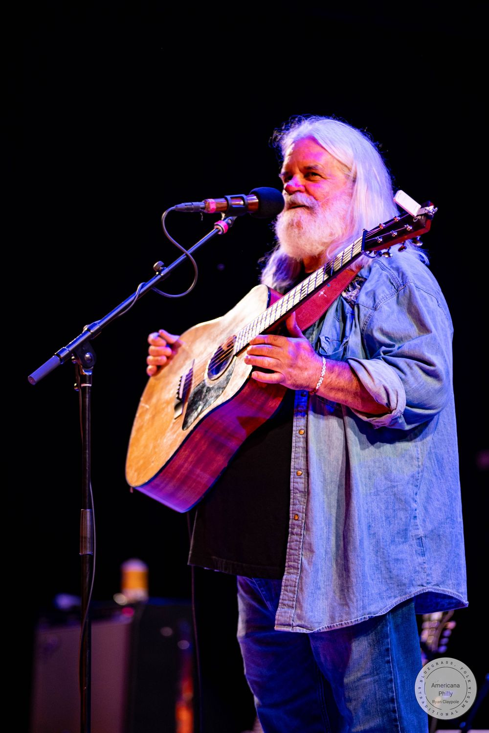 Elderly musician with long white hair and beard playing acoustic guitar and singing into microphone on stage.