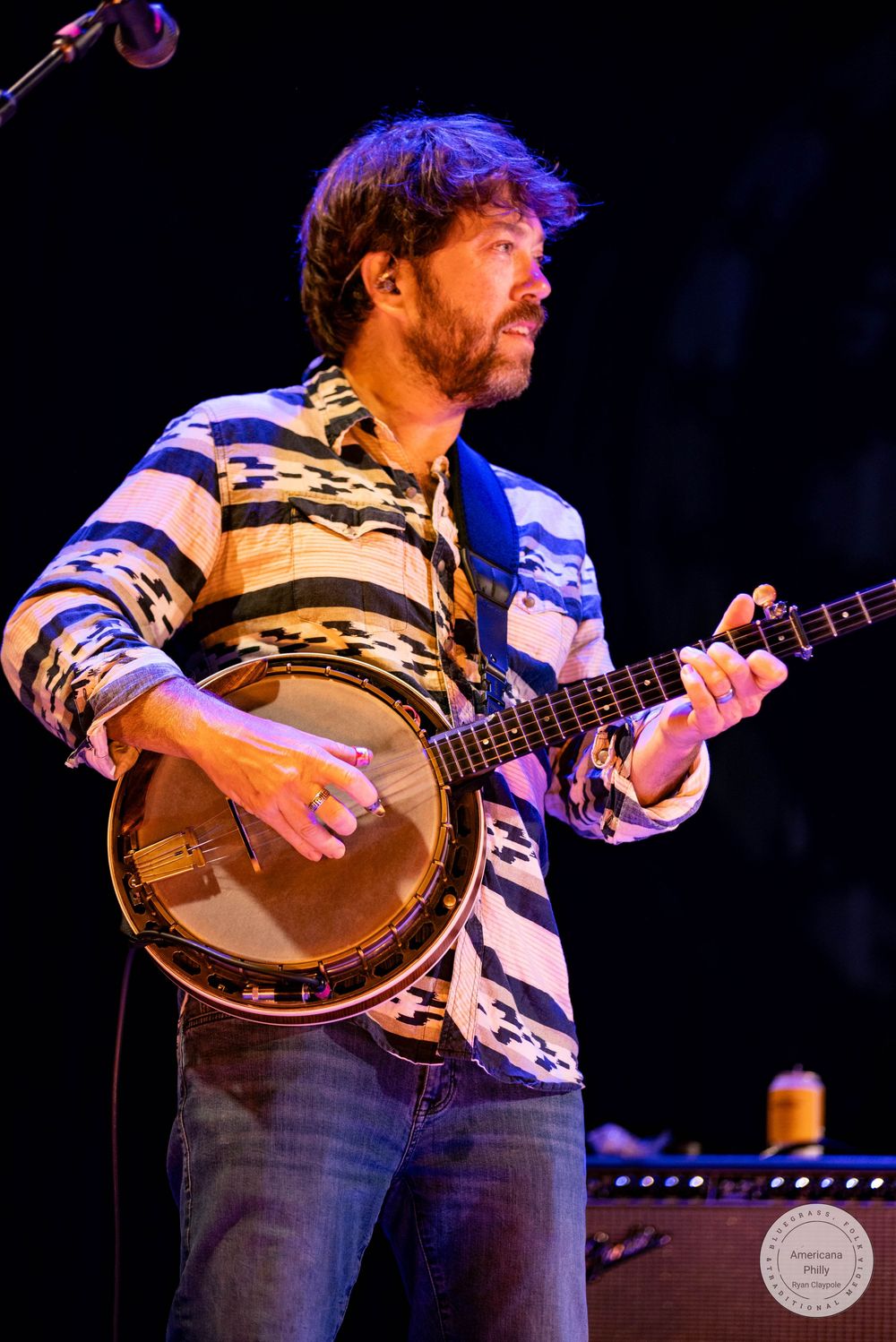 Man in patterned shirt playing banjo on stage under blue stage lighting.