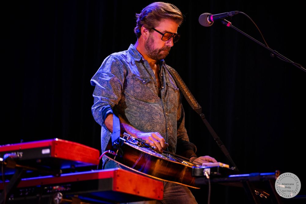 Bearded musician playing a lap steel guitar on stage with keyboards and a microphone at a live performance.
