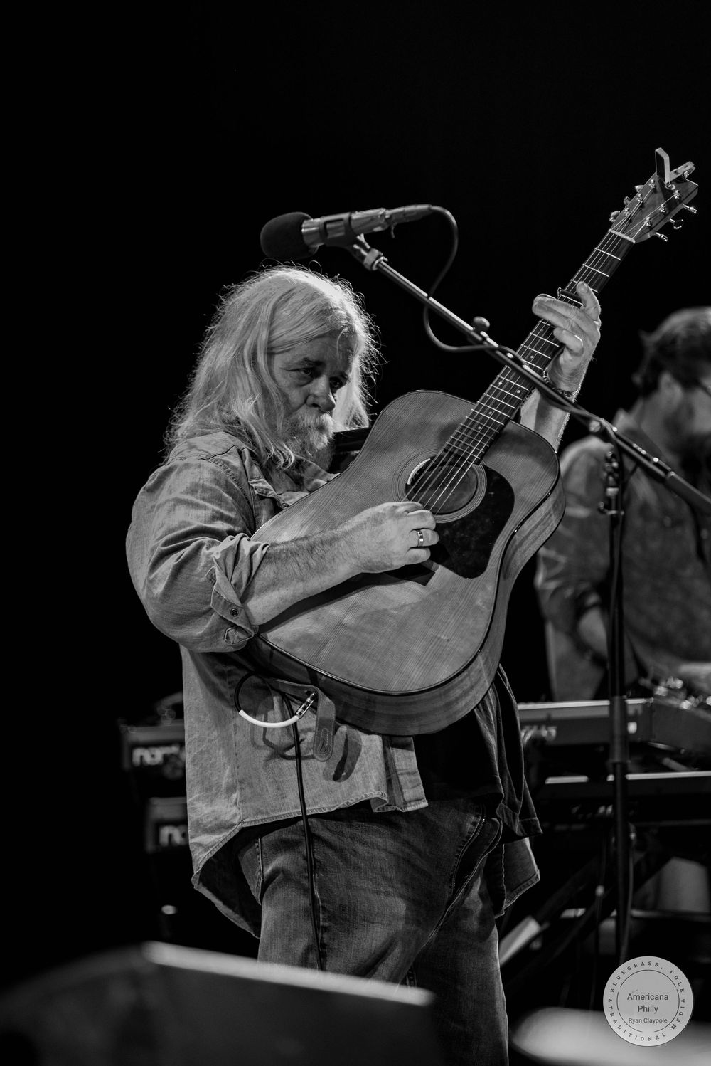 Black and white photo of a long-haired musician passionately playing an acoustic guitar on stage.