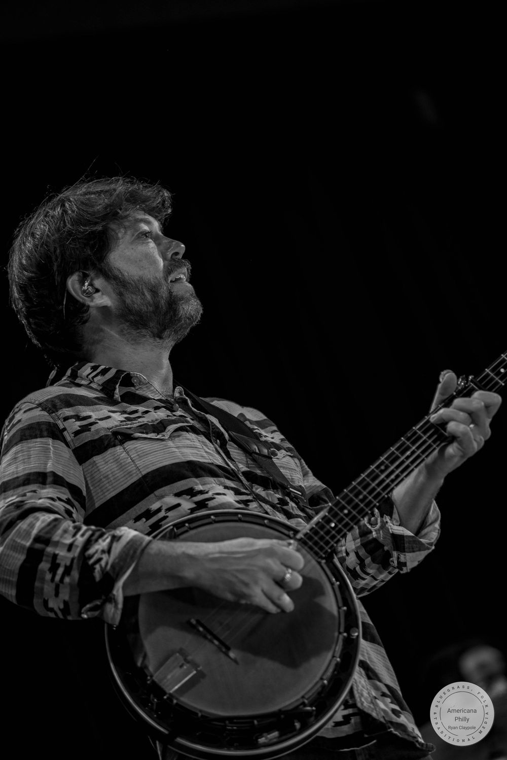 Black and white photo of a bearded man playing a banjo, looking upward, wearing a plaid shirt on stage.