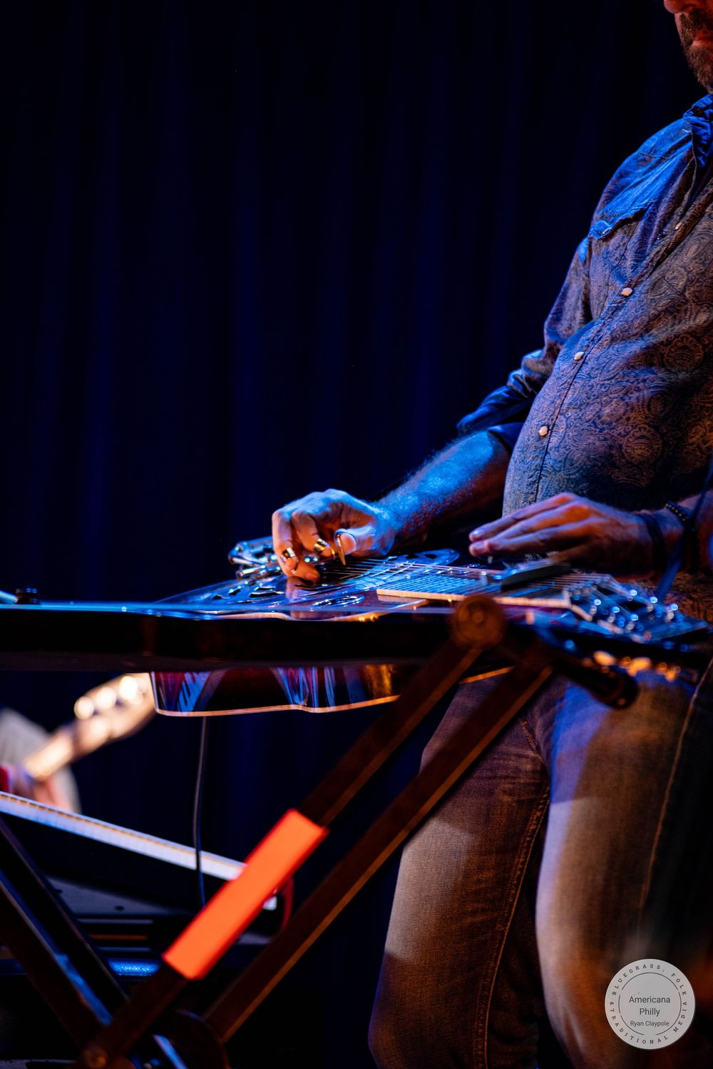 Musician playing a steel guitar on a dimly lit stage with blue and orange lighting.