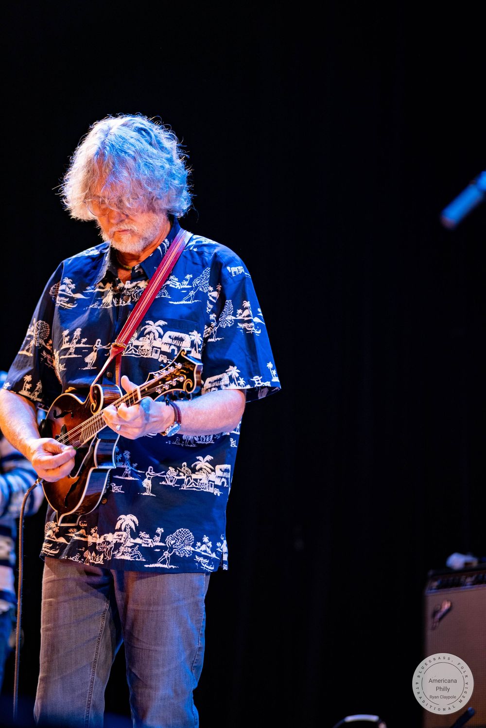Older musician with gray hair plays mandolin on stage wearing a blue Hawaiian shirt under dramatic lighting.