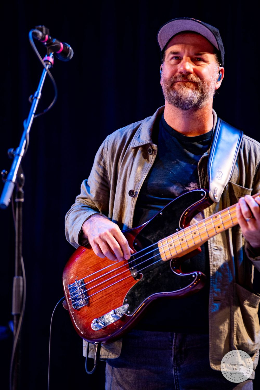 Smiling bearded bassist wearing a cap performs on stage with a red electric bass guitar.