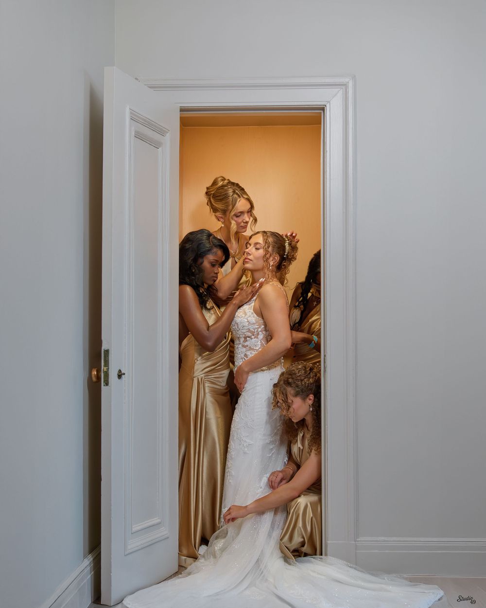 Bride in a white lace wedding gown is helped by four bridesmaids in gold satin dresses inside a doorway against a warm orange wall.