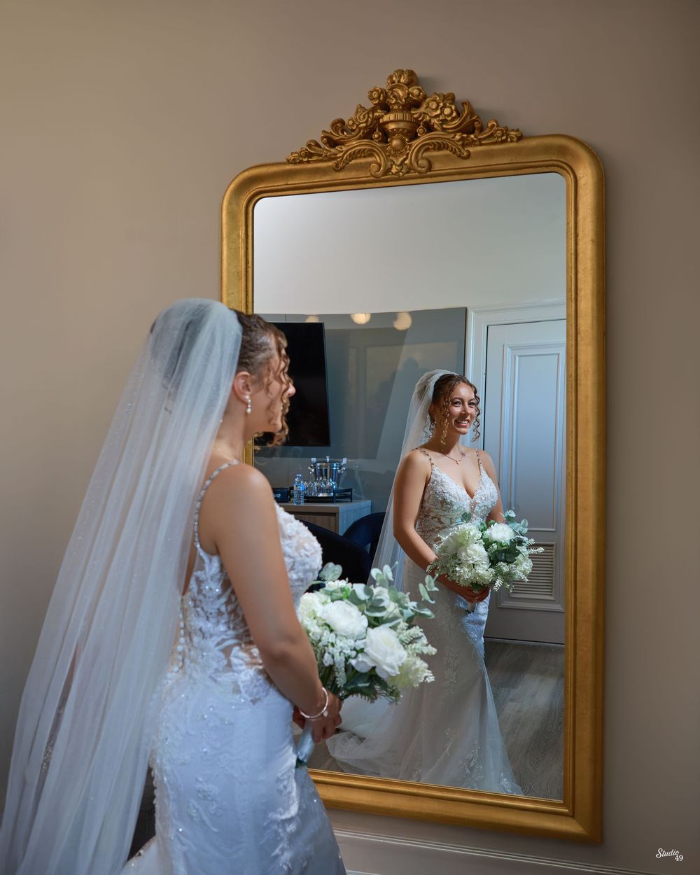 A bride in a lace gown and long veil holds a white floral bouquet and smiles at her reflection in a gold ornate mirror.