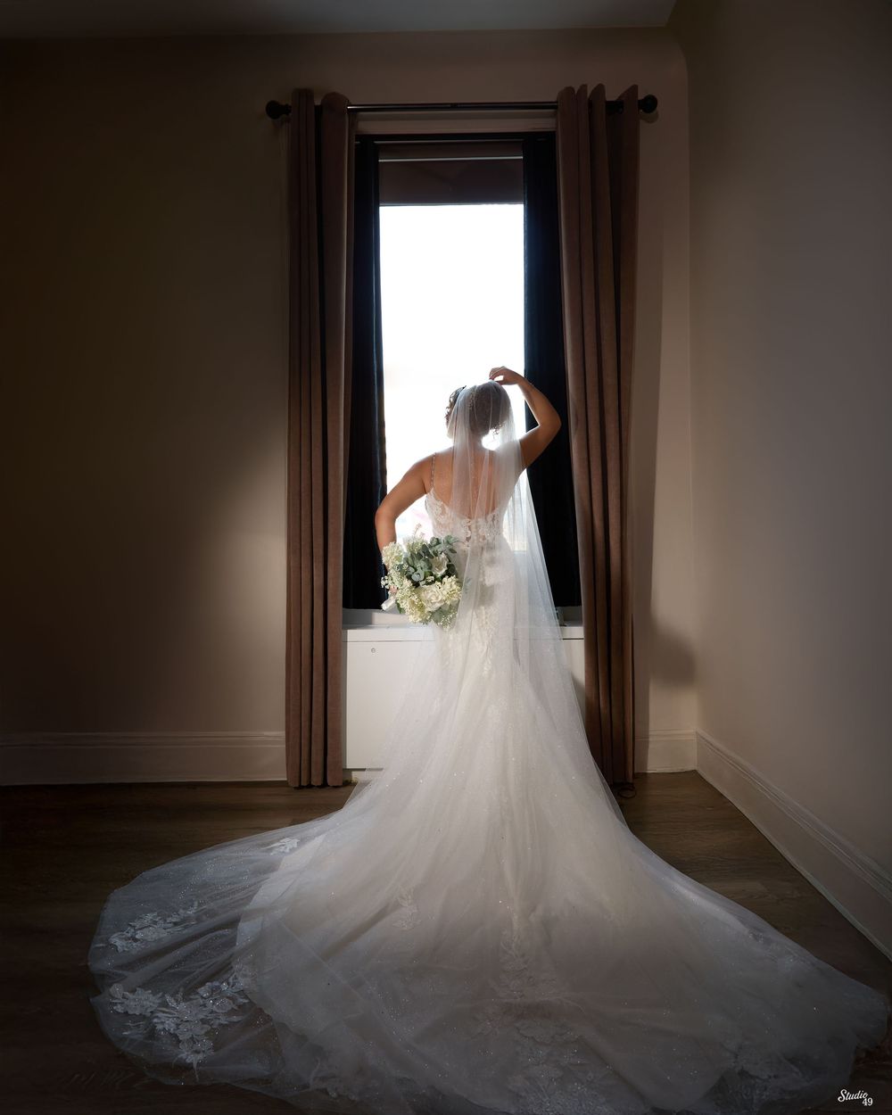 Bride in a flowing lace gown and long veil stands at a tall window, holding a white bouquet and touching her hair in soft natural light.