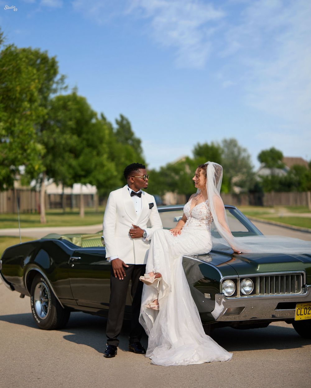 Bride in a white lace gown and veil sits on a vintage green convertible beside her groom in a white tuxedo jacket and bow tie.