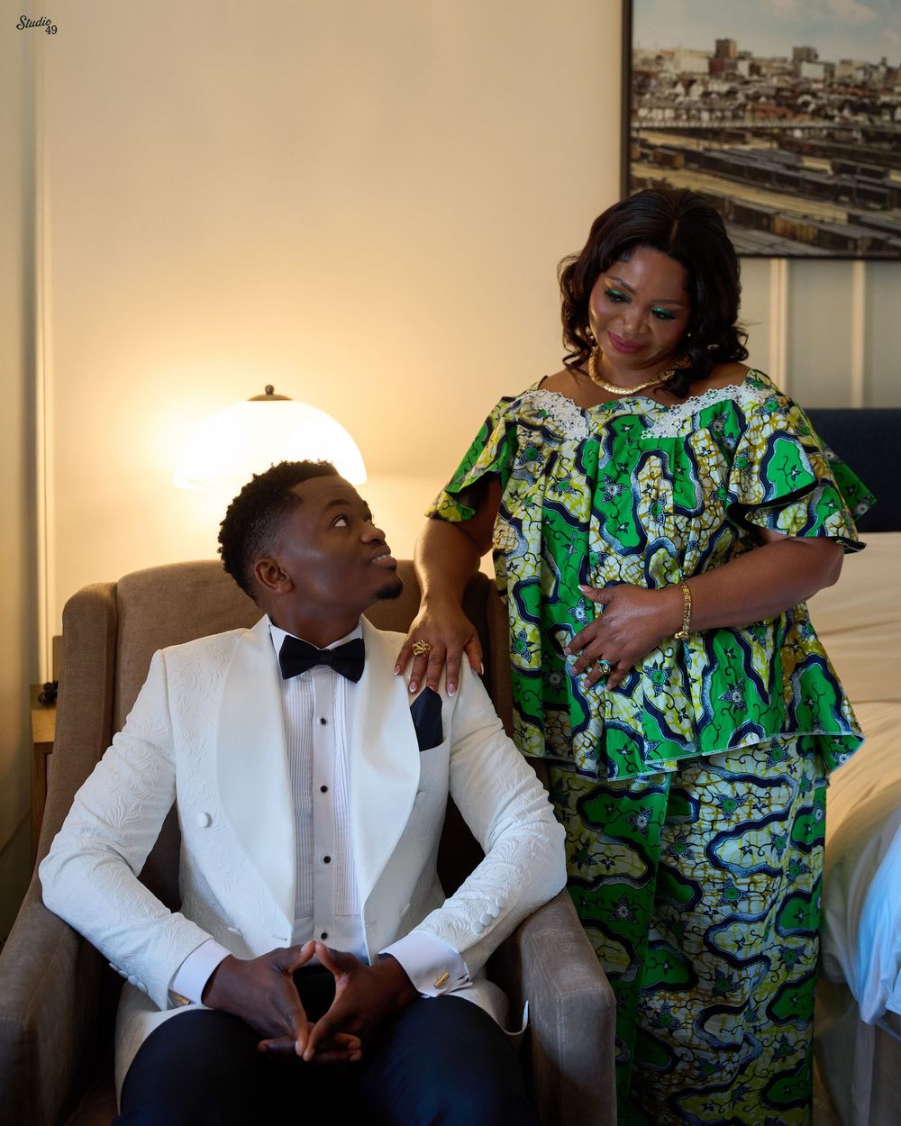 Groom in a white tuxedo jacket and black bow tie sits in an armchair while his mother in a green African print dress stands beside him.