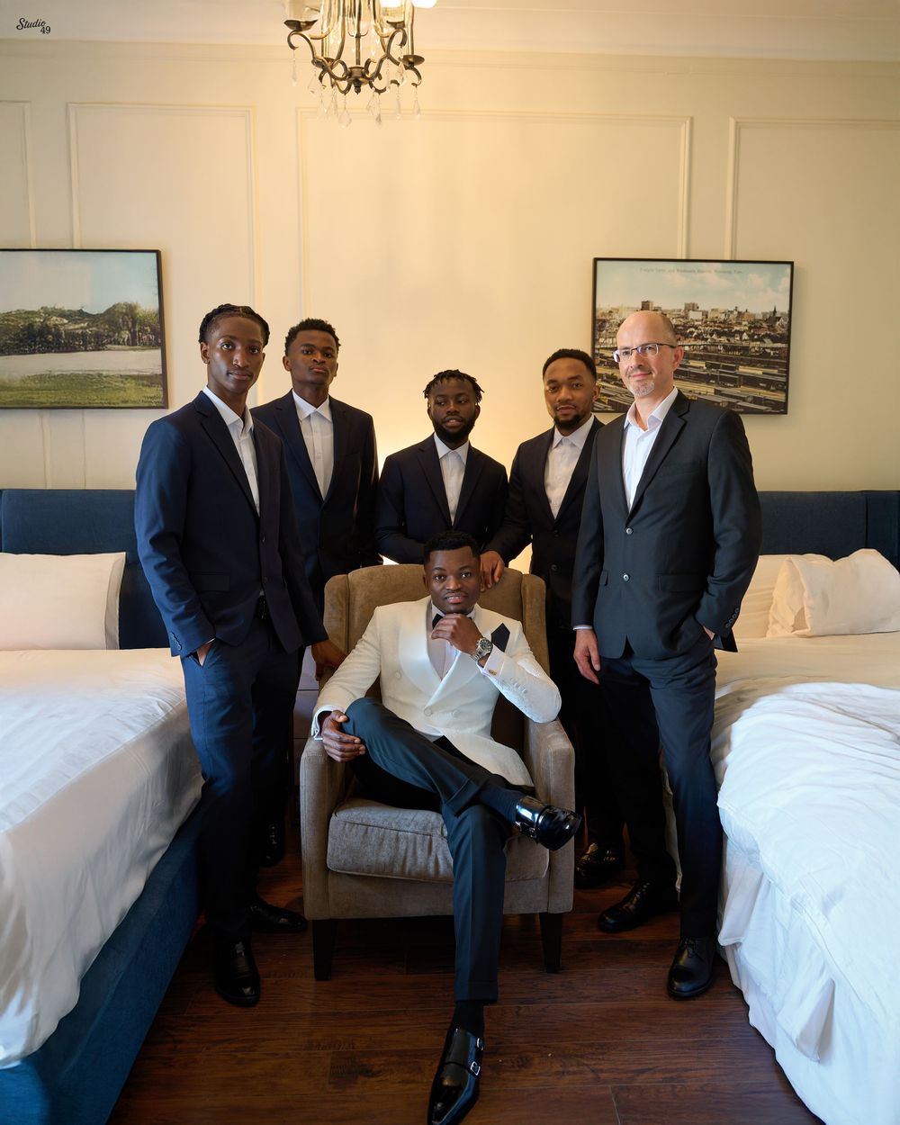 Five men in tailored dark suits stand around a seated man in a white blazer in an elegant hotel room with two beds and a chandelier.