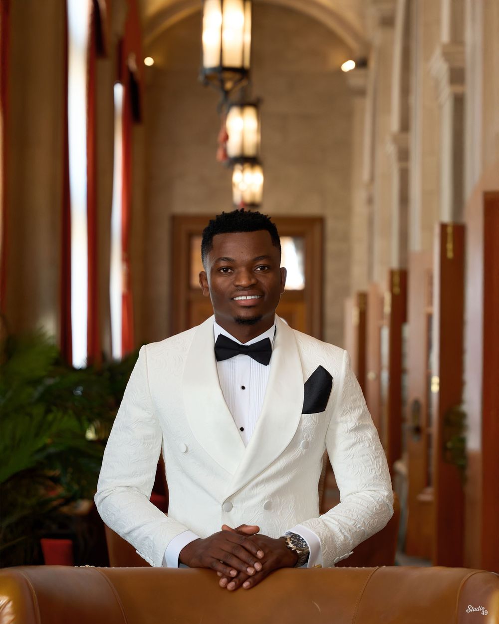 Smiling man in a white tuxedo jacket and black bow tie rests his hands on a leather chair in an elegant hall with tall windows.