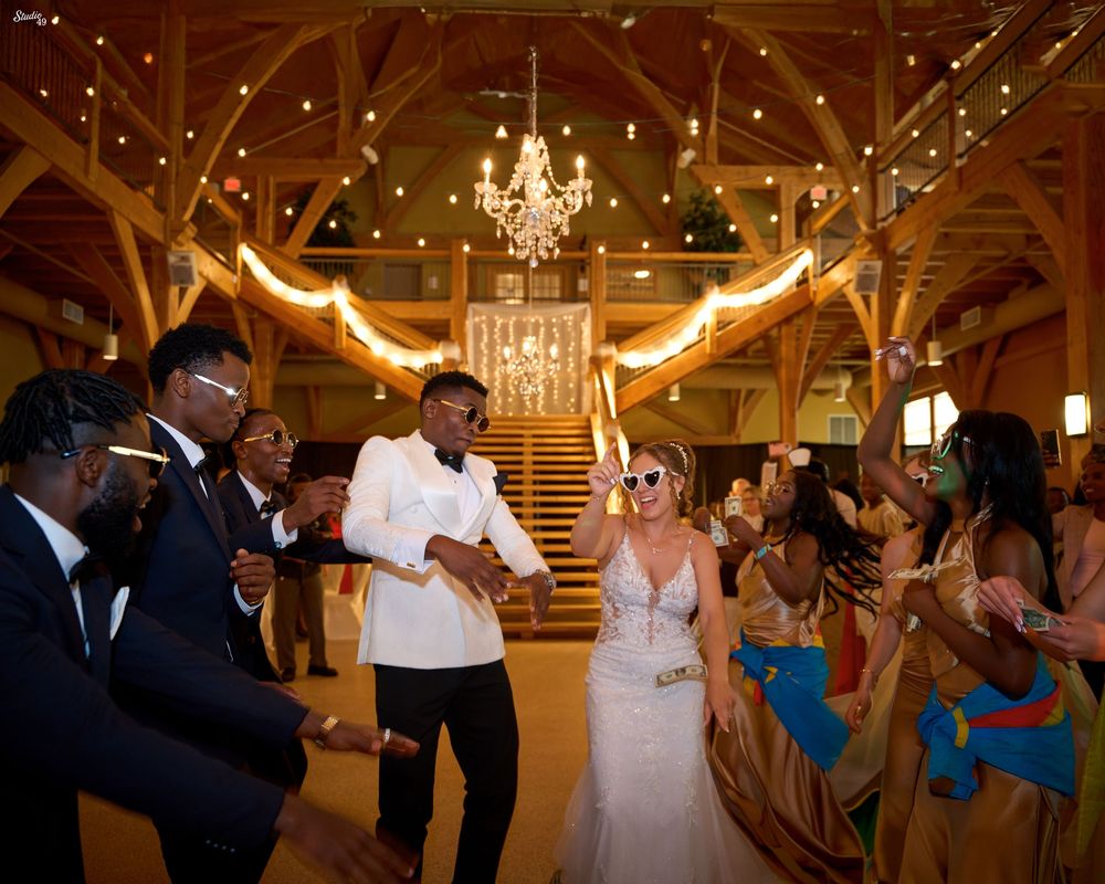 Bride in a white gown and groom in a white jacket dance with joyful wedding guests inside a rustic wooden venue lit by a chandelier.