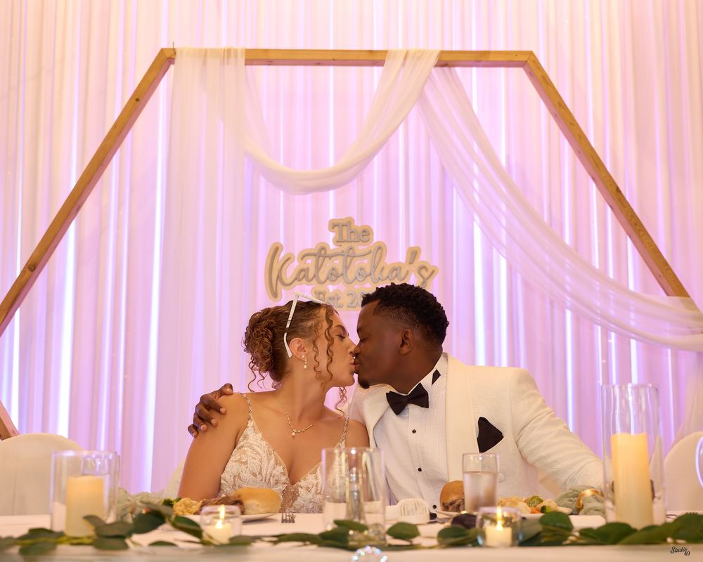Newlywed couple in wedding attire share a kiss at a sweetheart table with a hexagon backdrop, draped white fabric, candles and greenery.