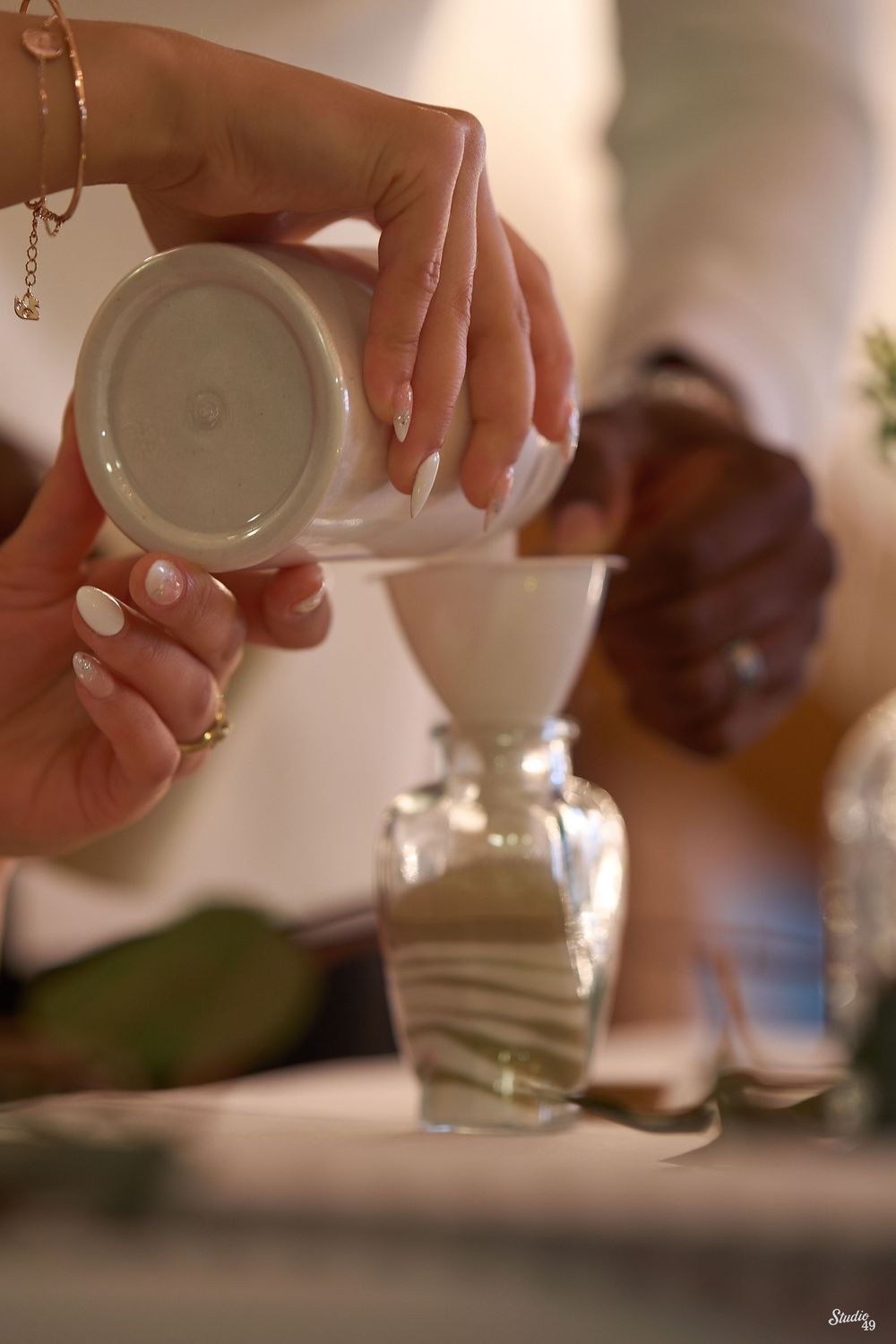 Hands pour white sand from a ceramic vessel through a funnel into a glass bottle of layered sand during a unity sand ceremony.