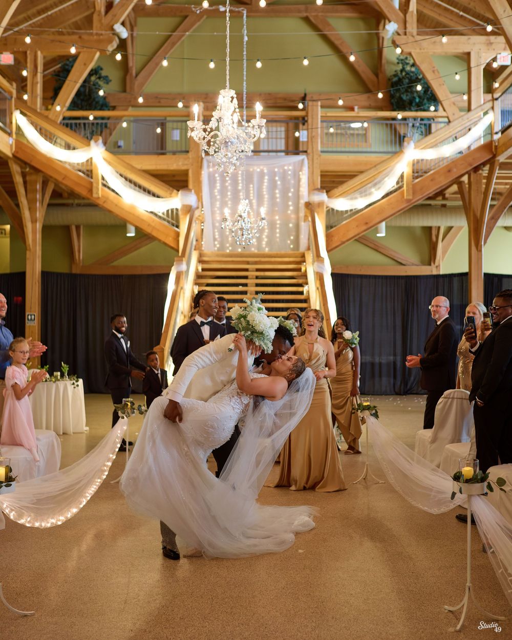 Groom dips bride in a romantic kiss at the end of the aisle as bridesmaids in gold gowns and guests watch in a rustic wooden venue.