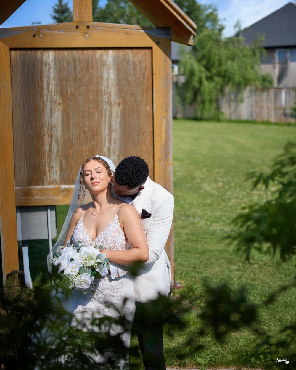 A bride in a lace gown and veil holds a white bouquet as her groom in a white jacket embraces her beside a rustic wooden structure.