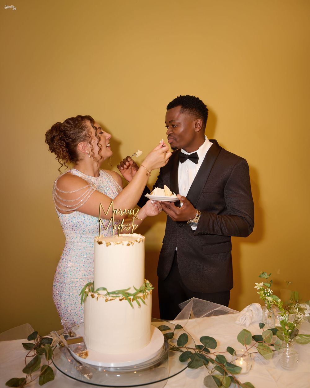 Bride in a sparkling gown feeds a bite of wedding cake to the groom in a black tuxedo and bow tie beside a white floral cake.