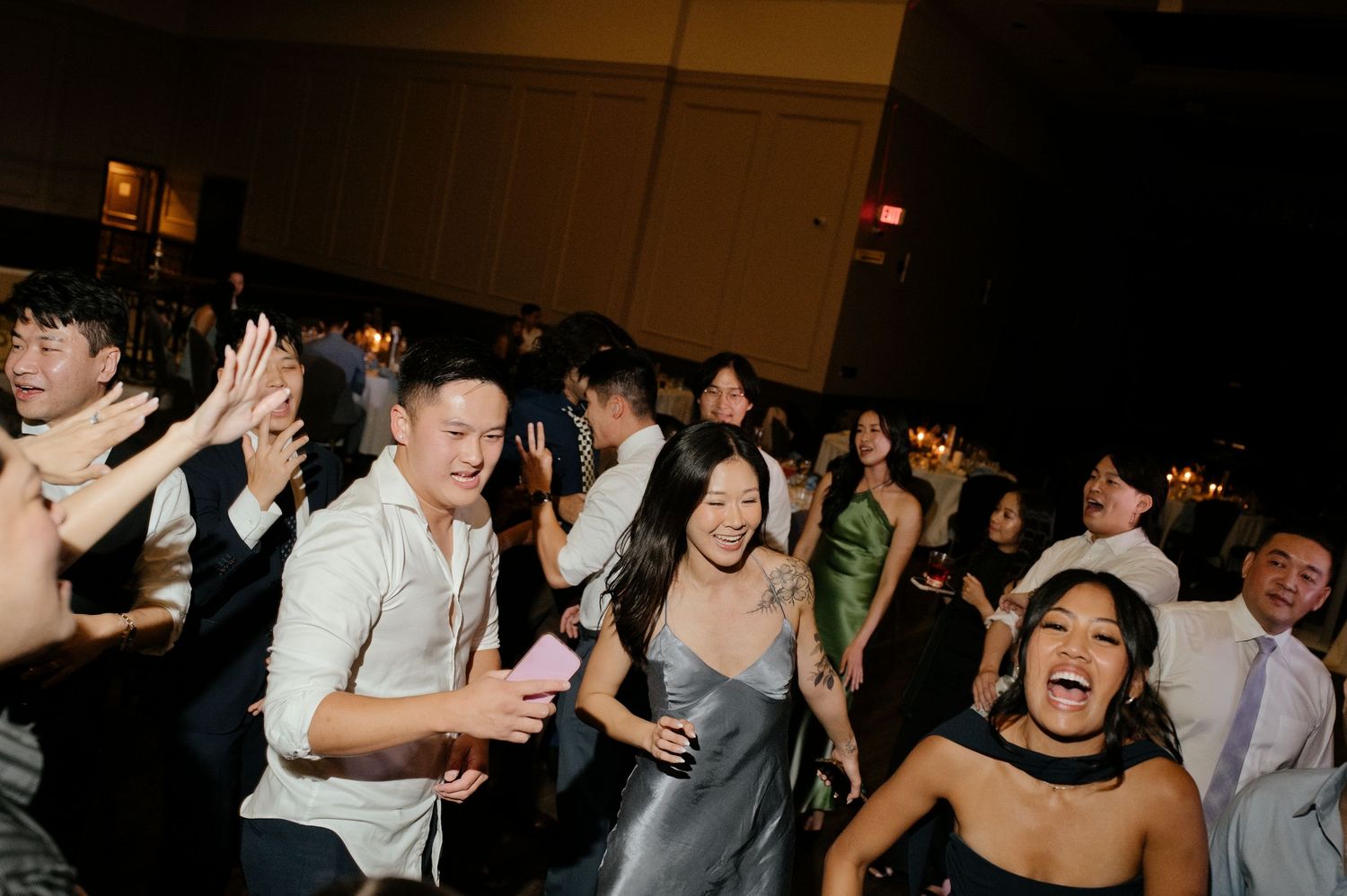 Guests in formal attire dance and laugh together on a crowded dance floor at a lively evening wedding reception.