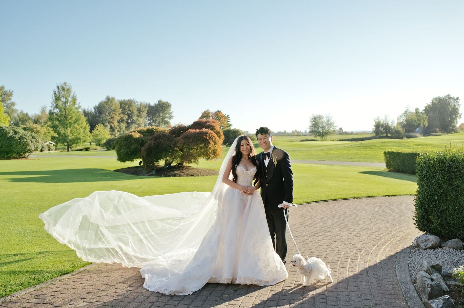Bride in a flowing white gown with a long veil and groom in a black tuxedo pose with a small white dog on a brick path by a golf course.