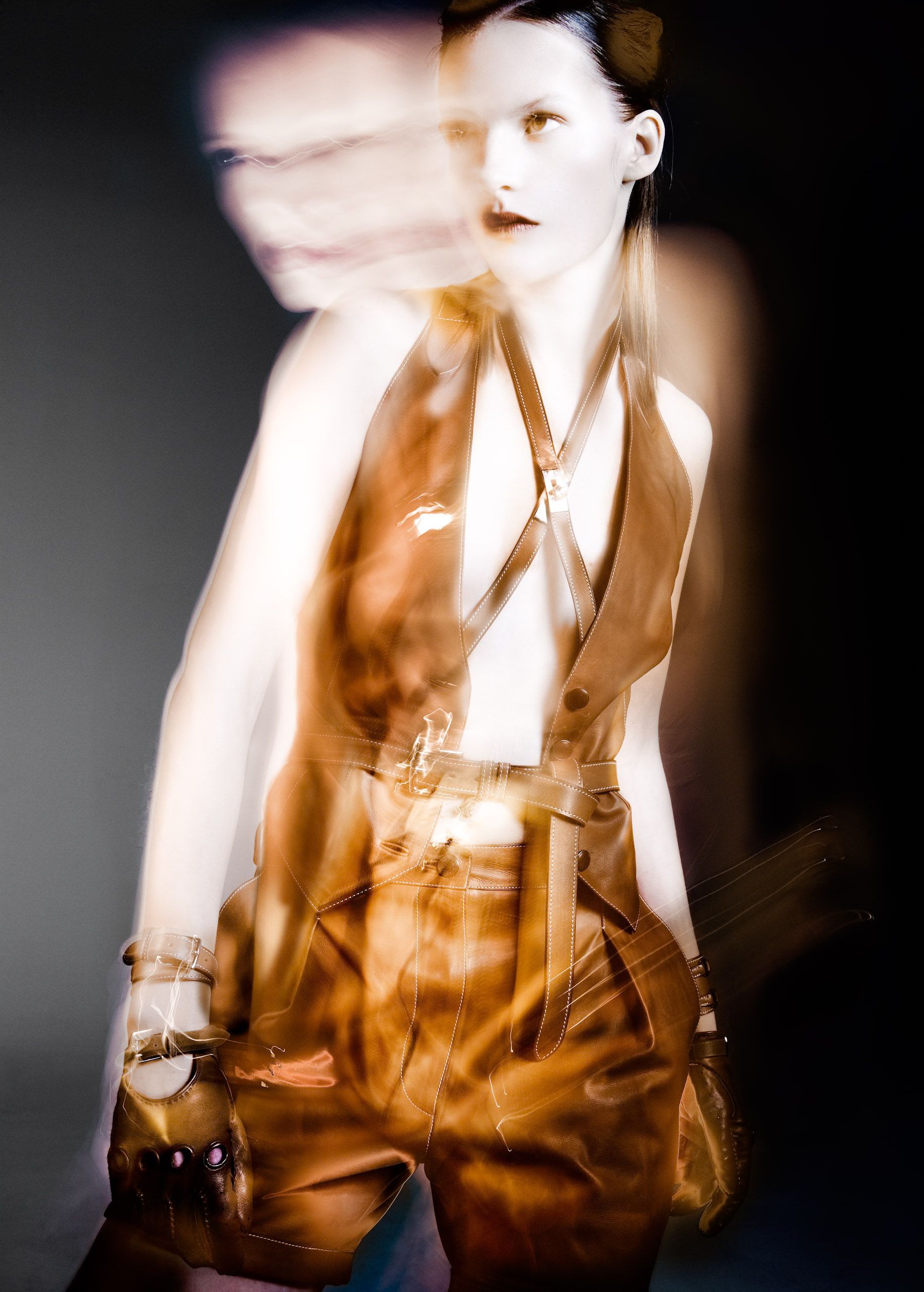 A model poses dynamically in brown leather harness top and shorts, captured with motion blur effect.
