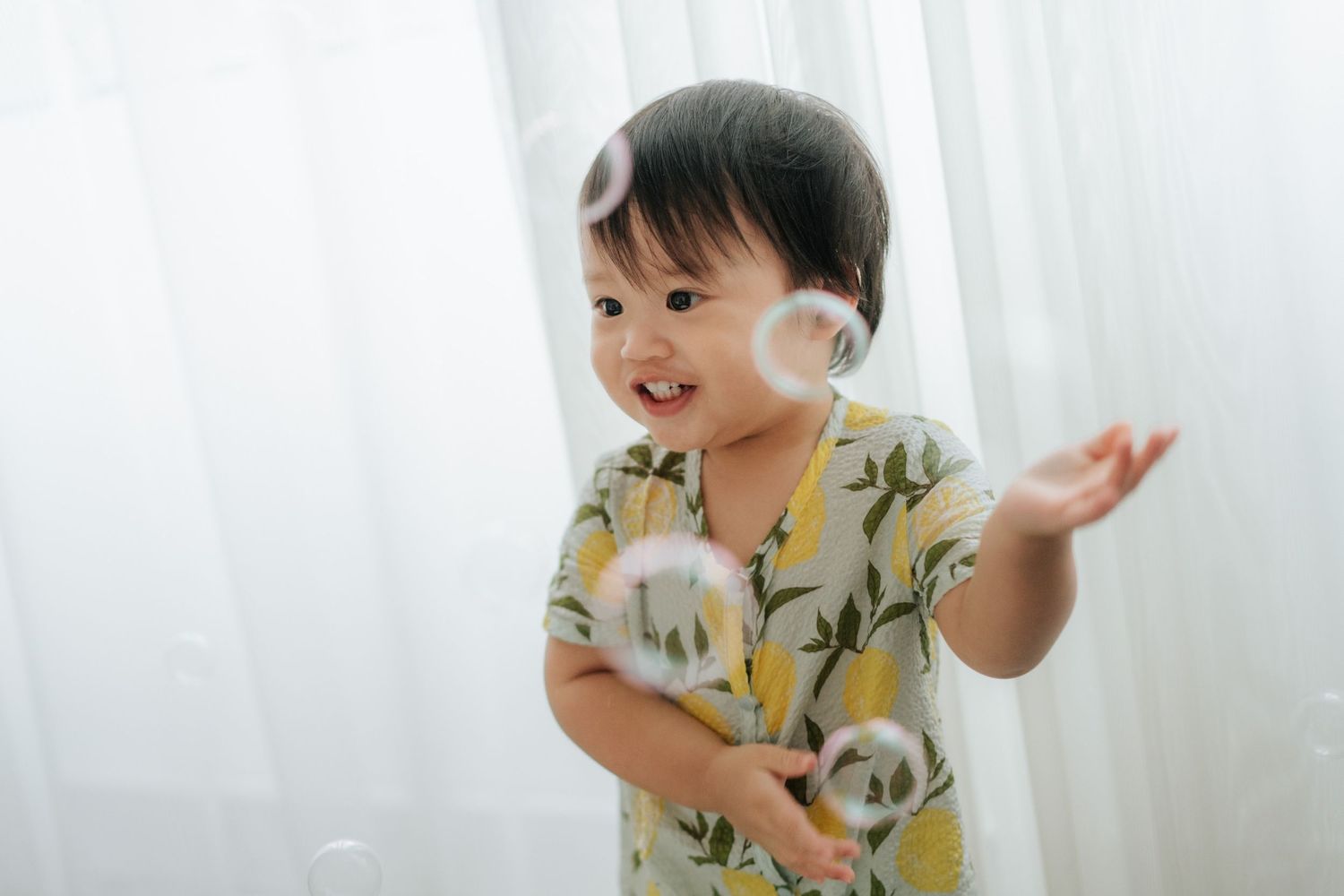 A happy toddler in a floral outfit reaches for floating soap bubbles indoors.