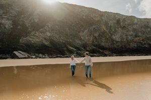 Couple holding hands walking on sandy beach with dramatic rocky cliffs and golden sunlight backdrop.