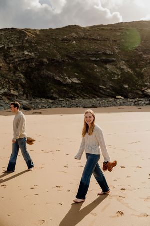 Couple walking barefoot on sandy beach holding shoes, wearing jeans and cream sweaters, with rocky cliffs in background.