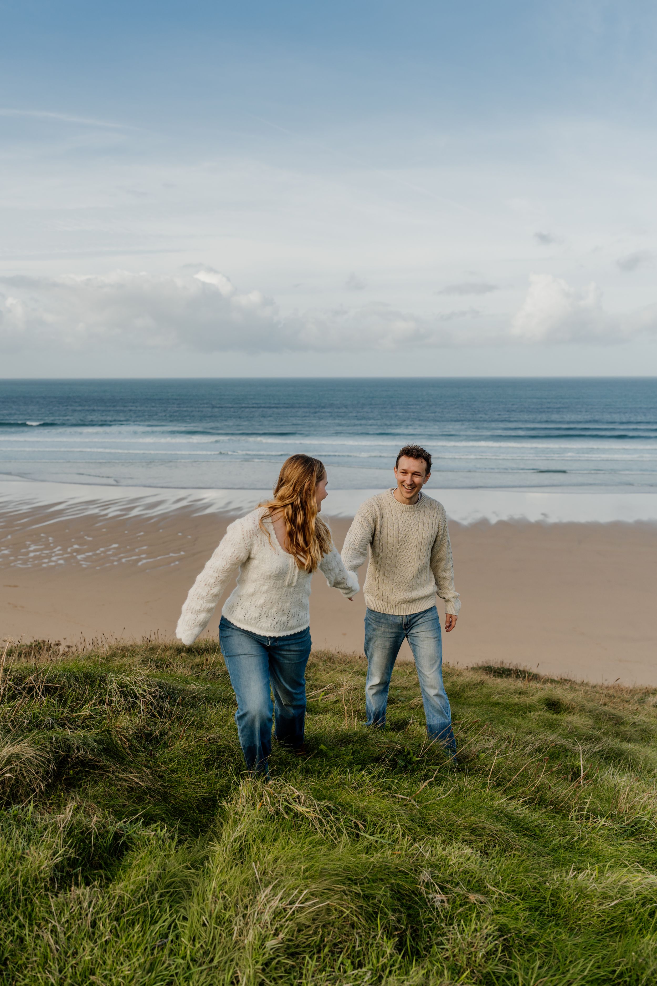 Couple holding hands on grassy coastal dunes with sandy beach and ocean in background.