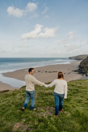 Couple holding hands on a grassy cliff overlooking a sandy beach and ocean under a partly cloudy sky.