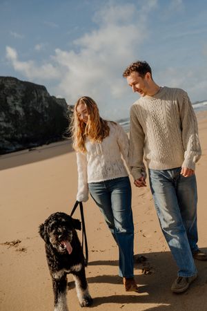 Couple in cable-knit sweaters walking a black dog on a sandy beach with cliffs in the background.