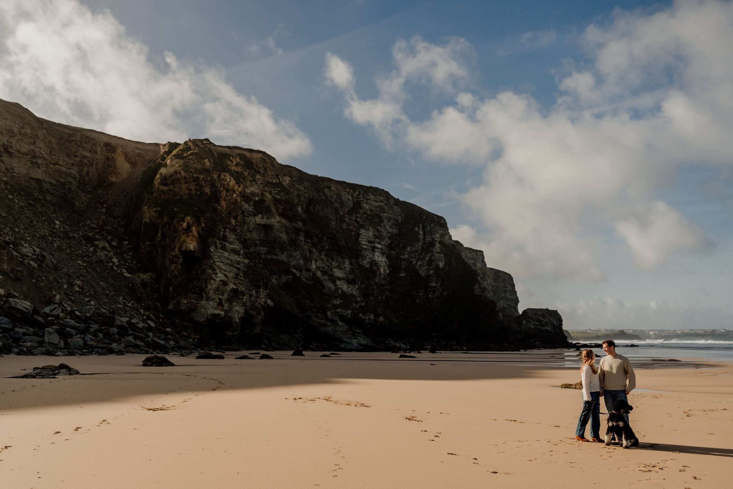 Two people walking on a sandy beach with dramatic rocky cliffs and ocean waves under a partly cloudy sky.