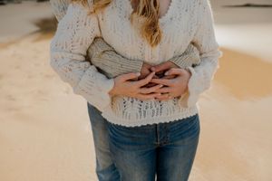 A woman in a cream knit sweater and jeans stands on a sandy beach with hands clasped over her stomach.