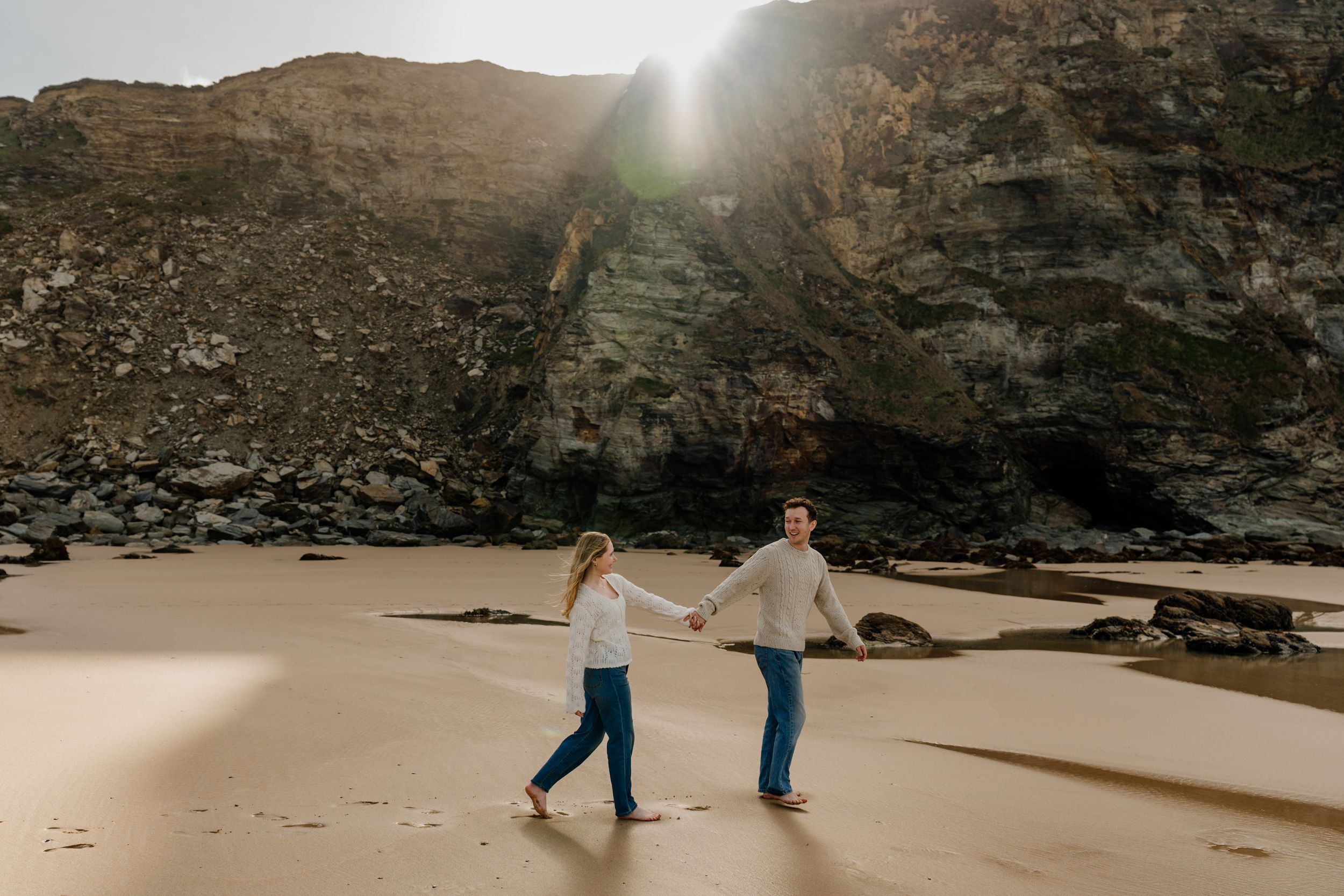 Couple holding hands walking on sandy beach with dramatic rocky cliffs and sunburst in background.