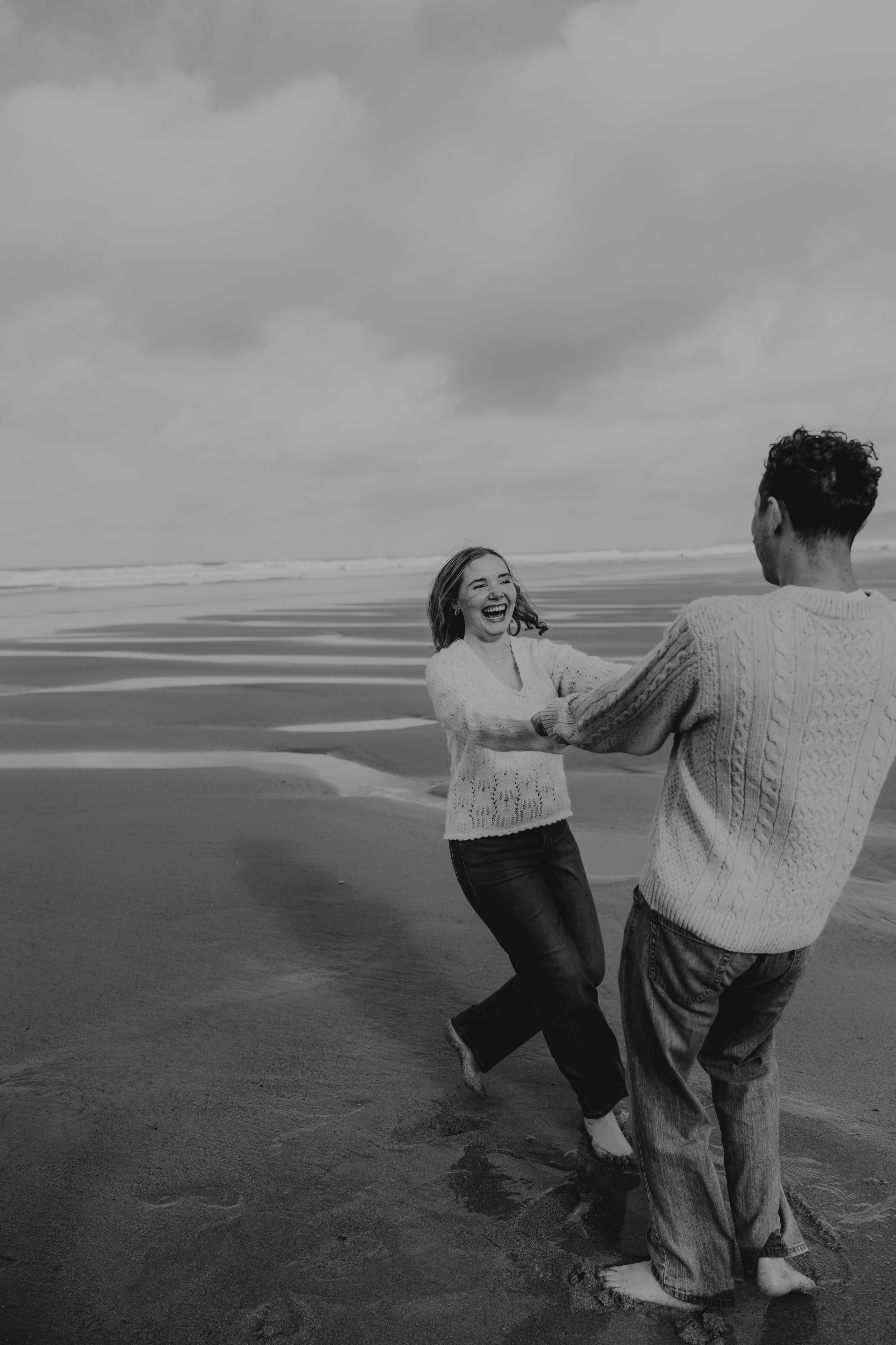 Couple dancing barefoot on a beach in black and white, woman laughing joyfully while holding hands with her partner.